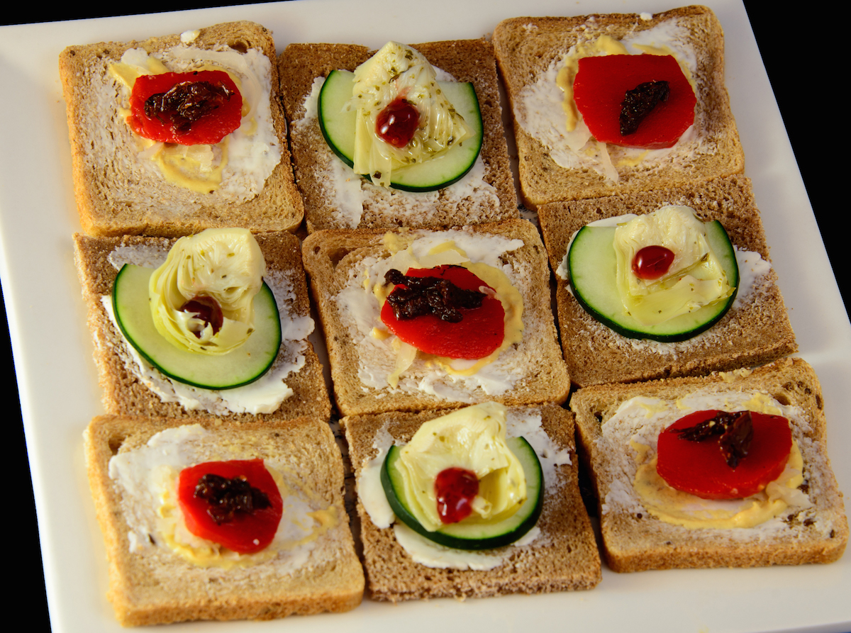 A plate of open face Danish-style sandwiches, as seen from above, arranged in a checkerboard pattern. These are the vegetarian sandwiches, featuring roasted red bell pepper cut into half round shapes, and pickled artichoke hearts cut into trapezoids.